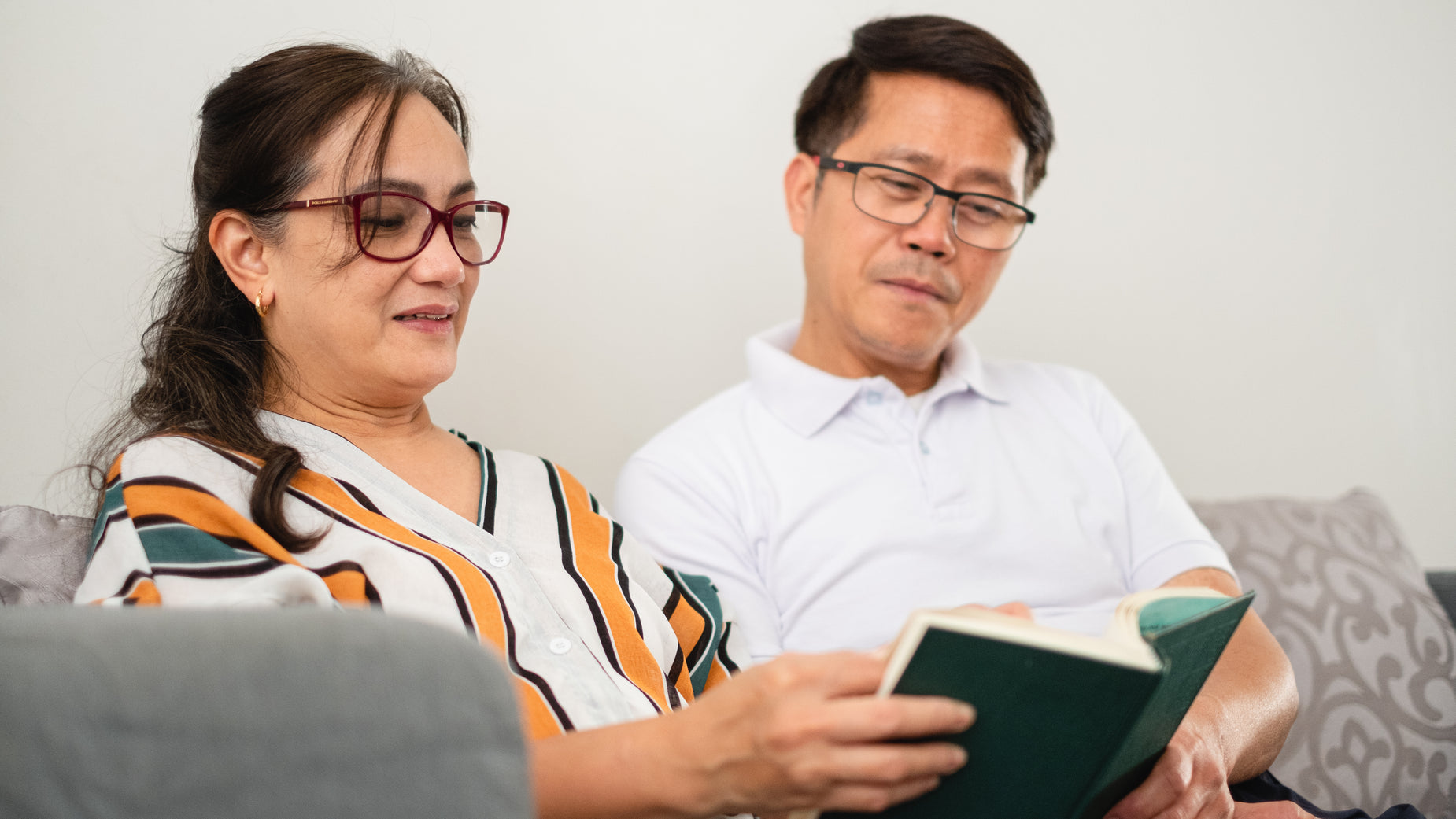 an asian man and woman sitting on a couch looking at a book