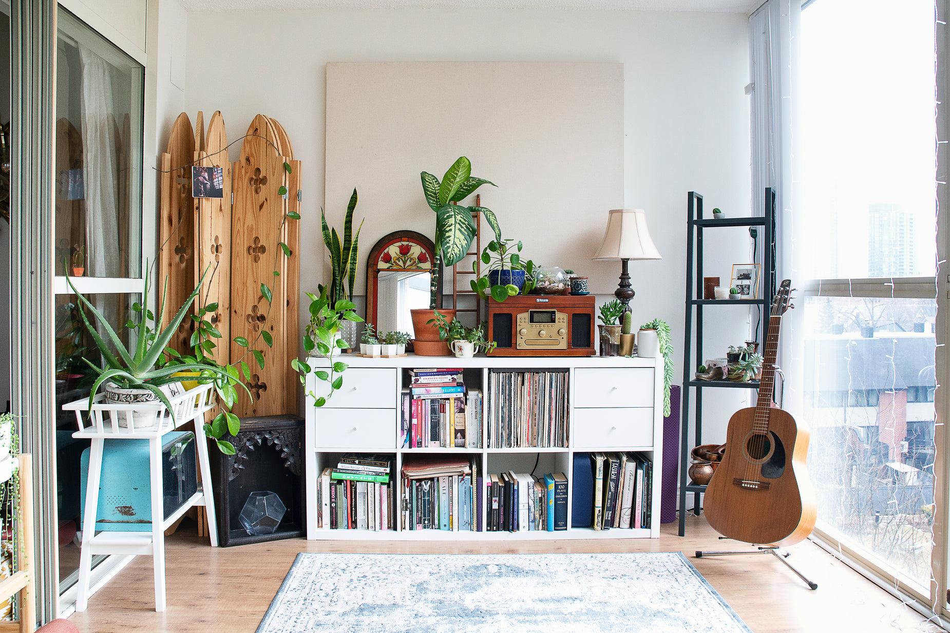 the living room is cluttered and has plants, bookshelf and a guitar
