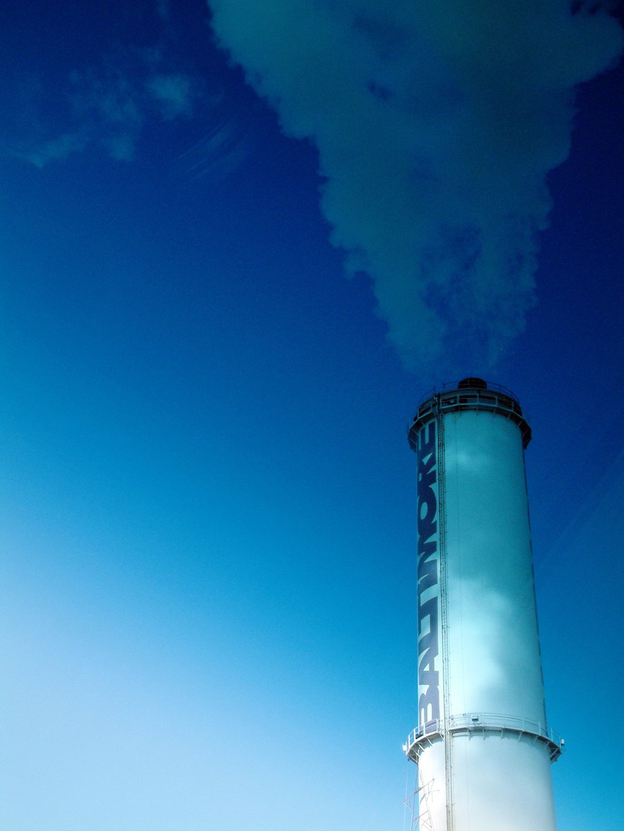smoke stack rises into the sky with blue water in front of it