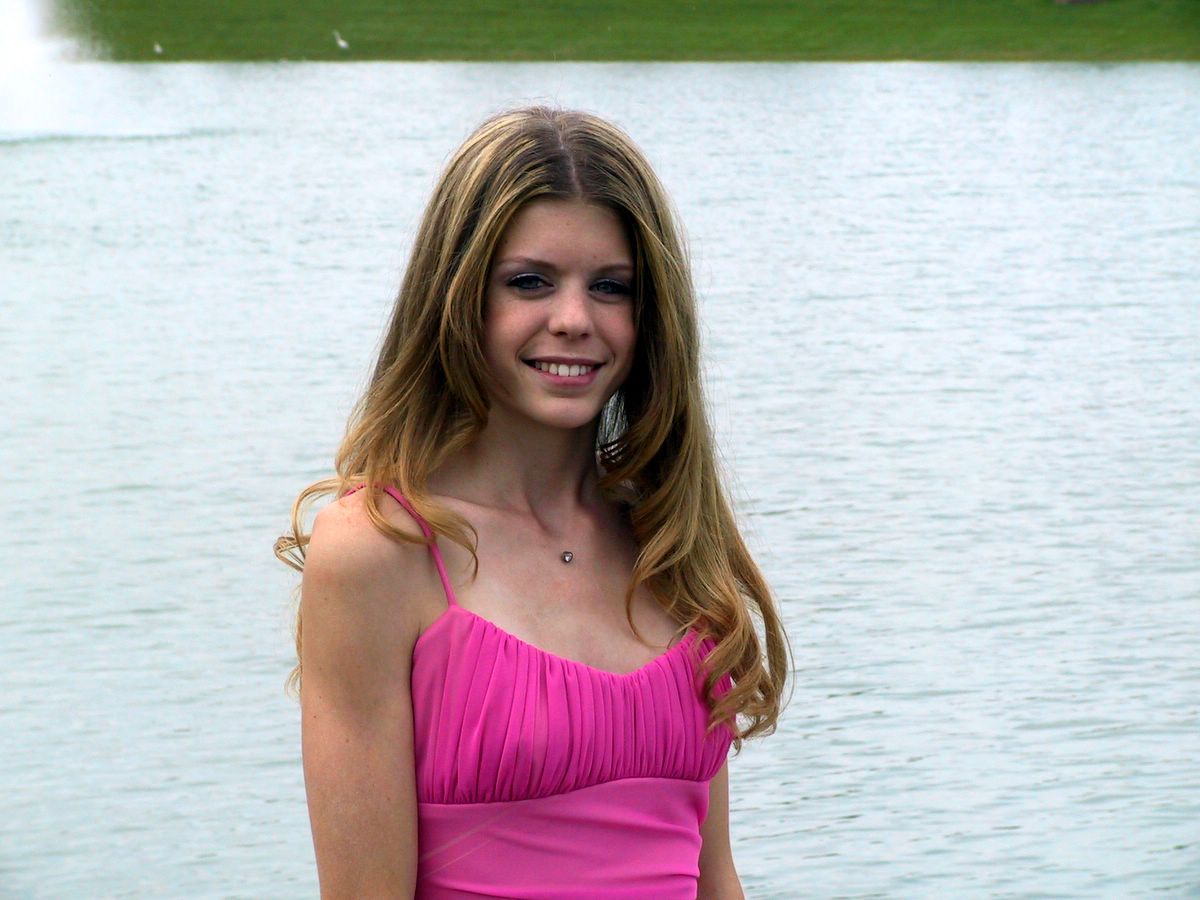 a smiling girl in a pink dress posing next to a lake