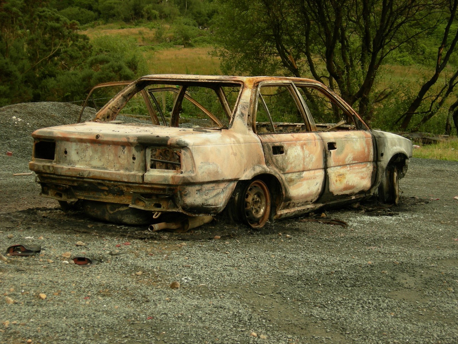an old, dirty car that has rusted, is parked on a gravel road with trees in the distance