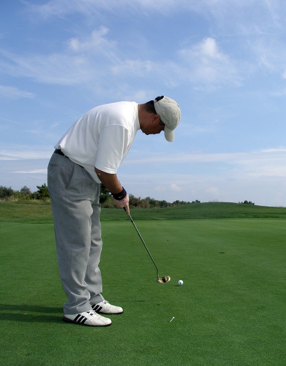 a man with white shoes and a cap is bent over golf ball and his arm is holding a club