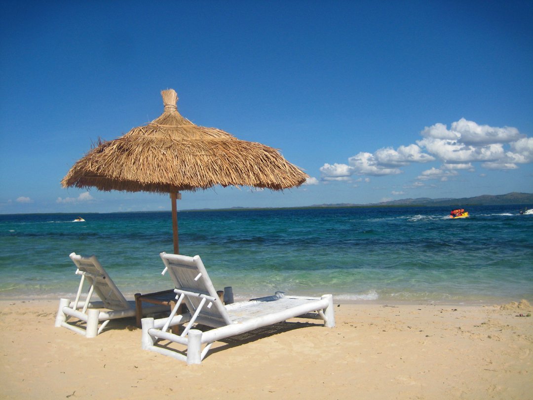 beach scene with two white lawn chairs, straw umbrella, and boats