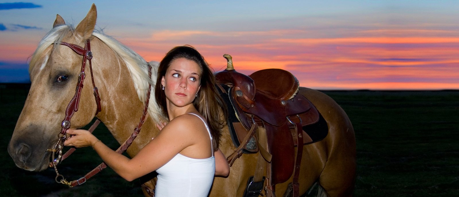 a pretty woman standing next to a horse at sunset