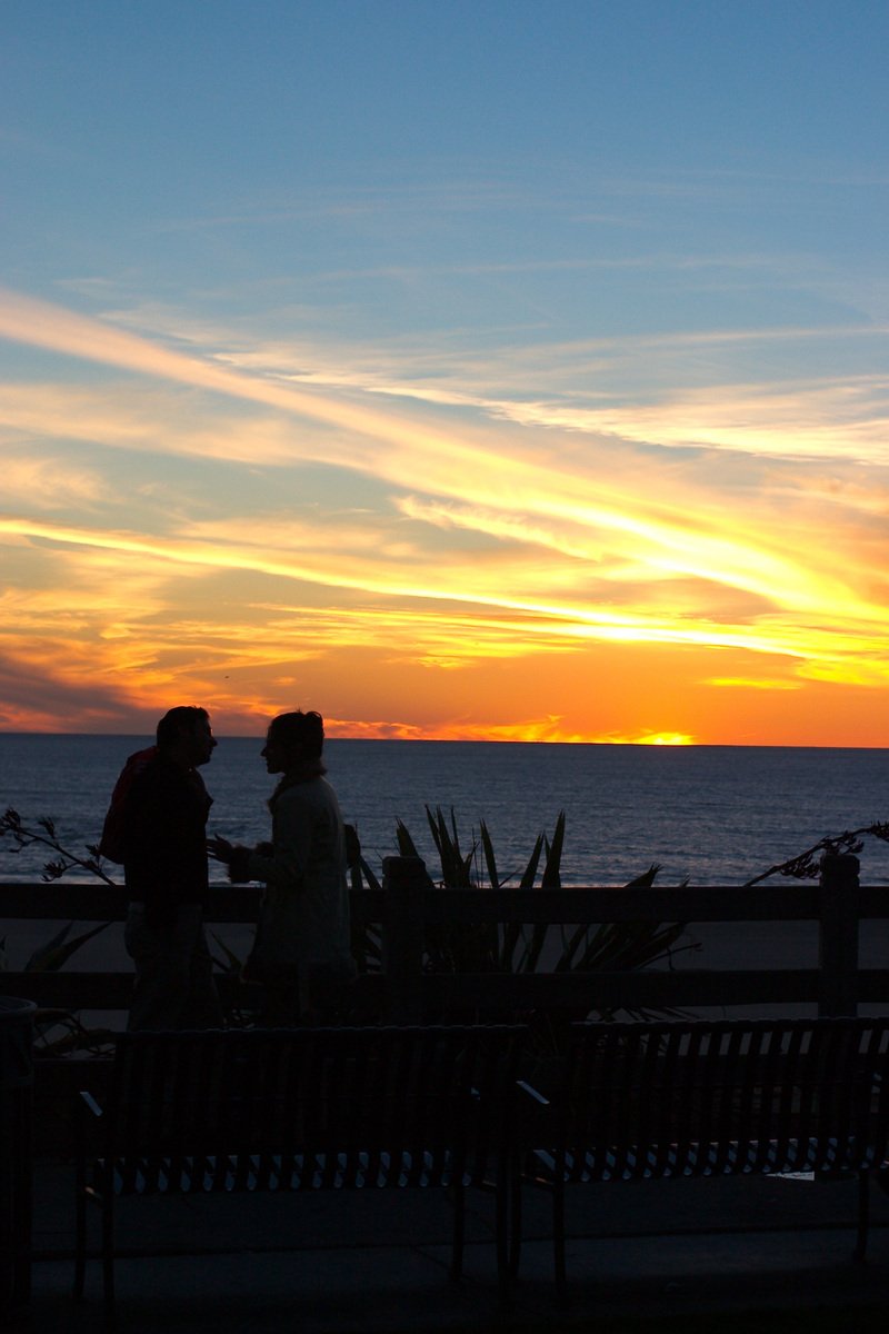 two people standing near a bench with a sunset