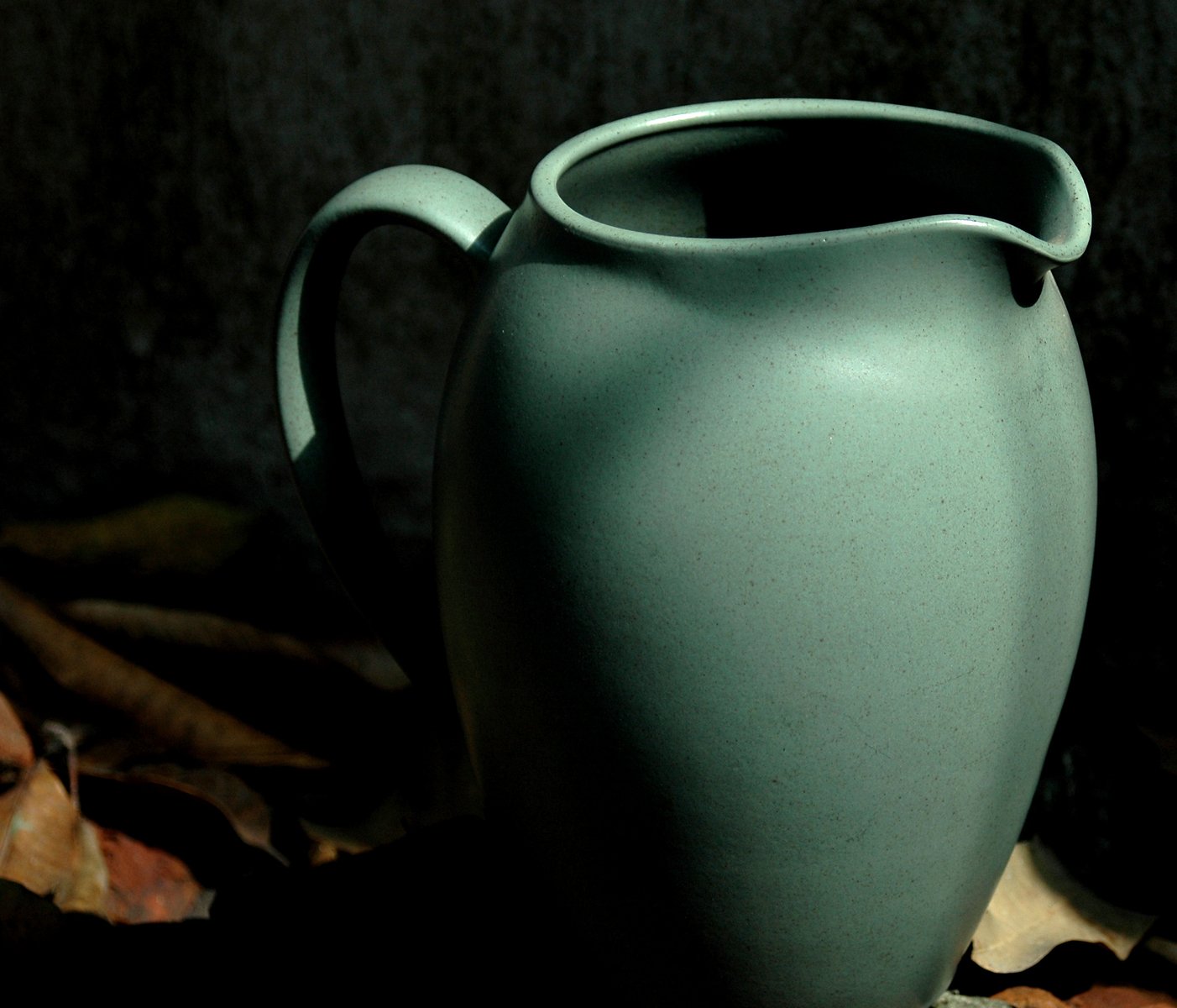 a green pitcher sitting on the ground with autumn leaves around it