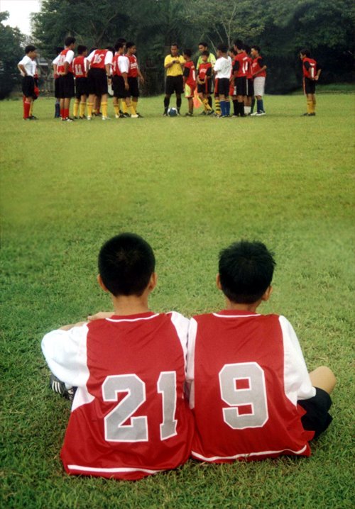 two boys sit in the grass as they watch a soccer team