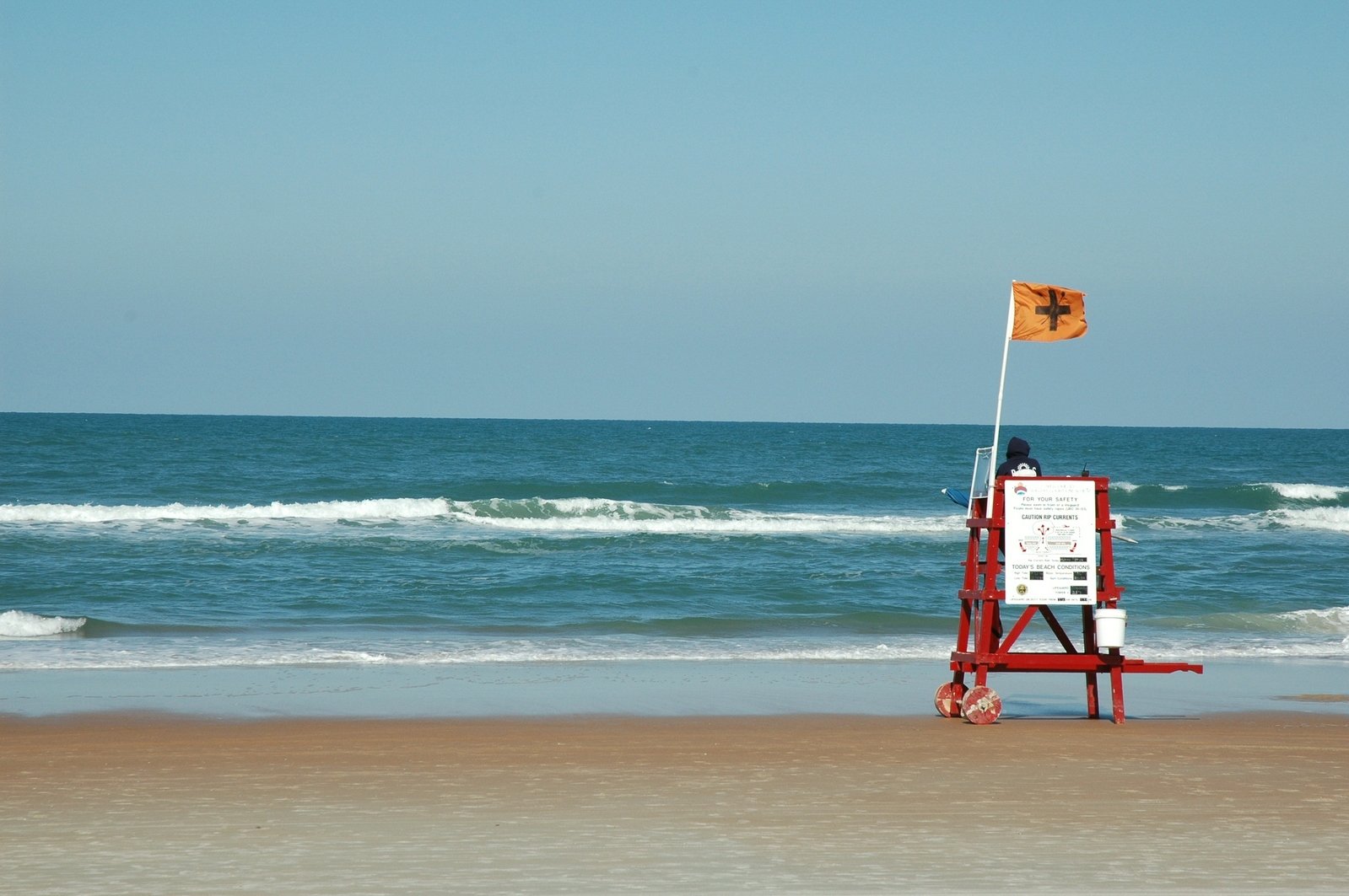 a white sign with a brown flag on top sitting in the sand near a beach