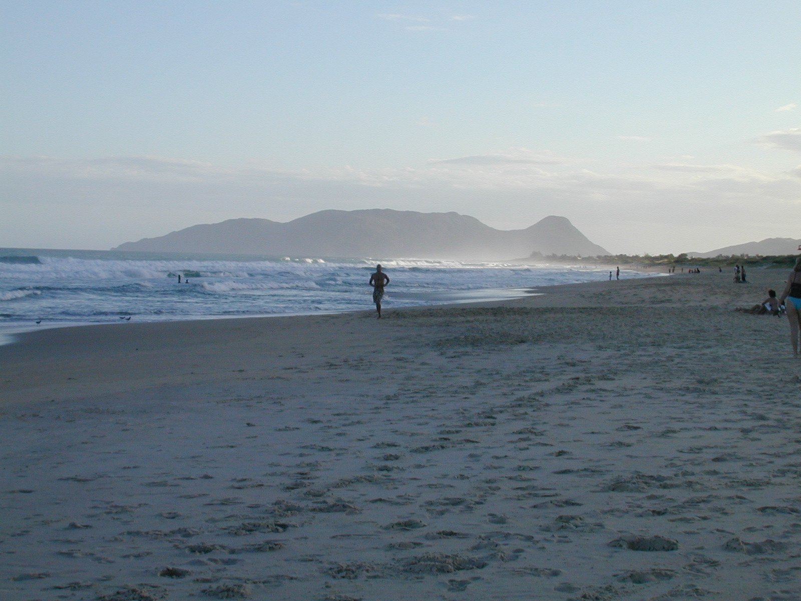 a beach with people walking in the sand and a person walking on the beach holding a surfboard