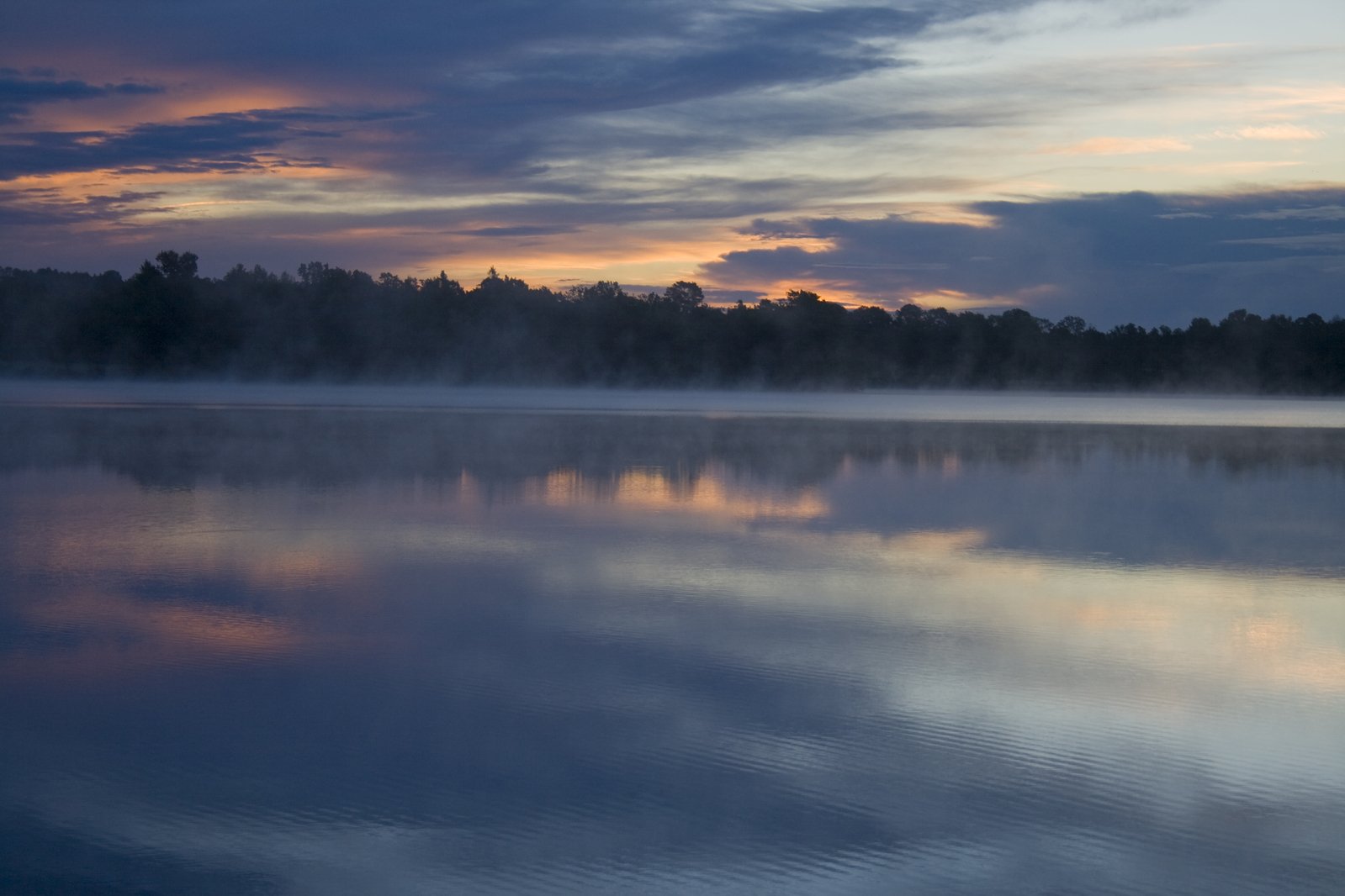 a foggy sunset shines over the calm water of a lake