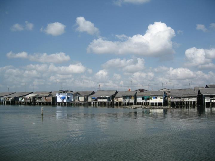 a river side neighborhood with a wooden pier, lots of buildings and a cloudy sky