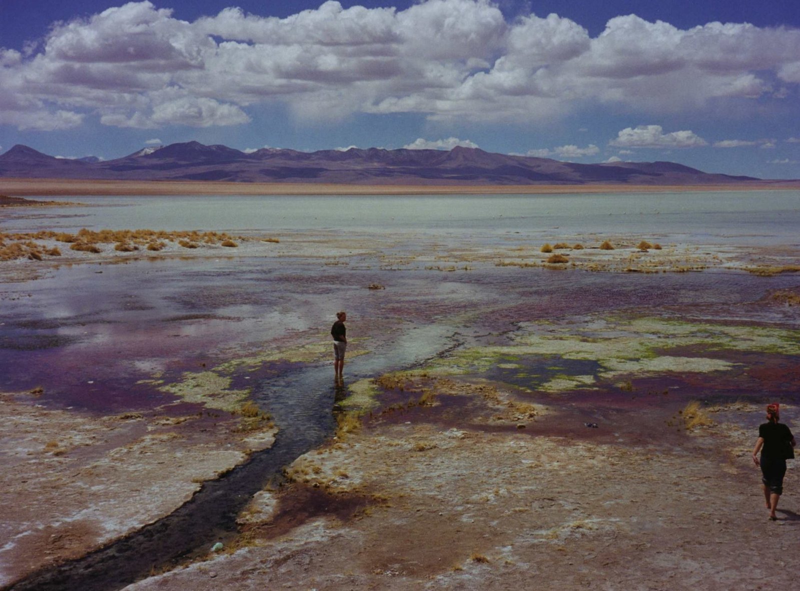 two people walking on a rocky area in front of mountains