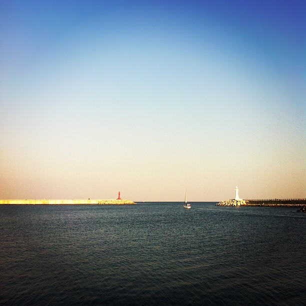 sail boats are on the water and a long bridge has a light tower in the distance