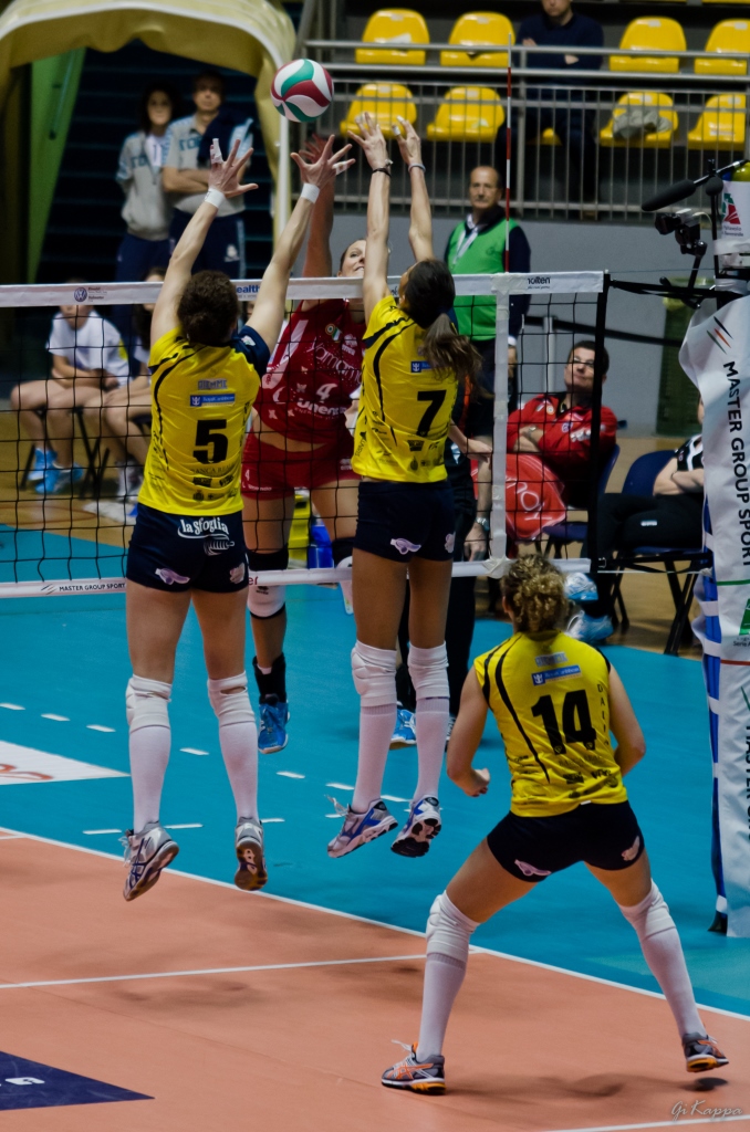 four volleyball players playing volleyball while one woman jumps up to block the ball