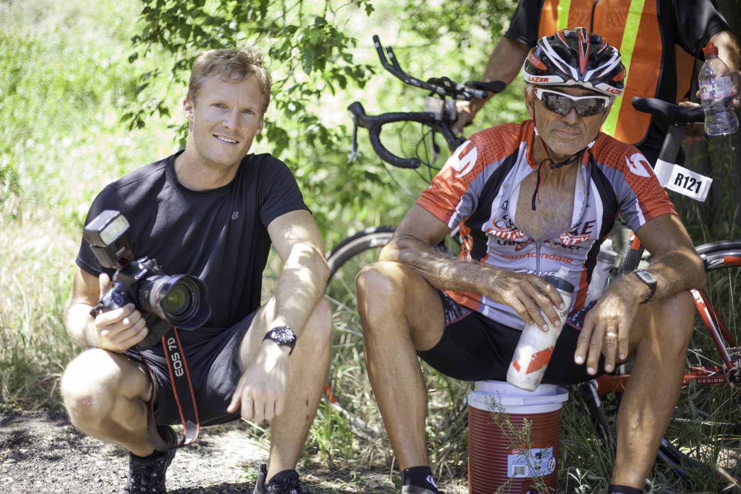 two men sit next to each other with bicycles in the background