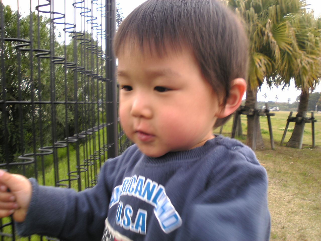 an infant boy in a blue shirt holds soing in front of a fence