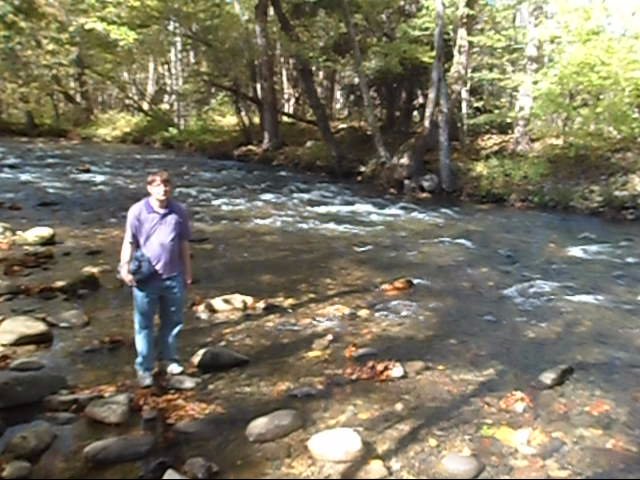 man standing next to rock in middle of creek with trees around