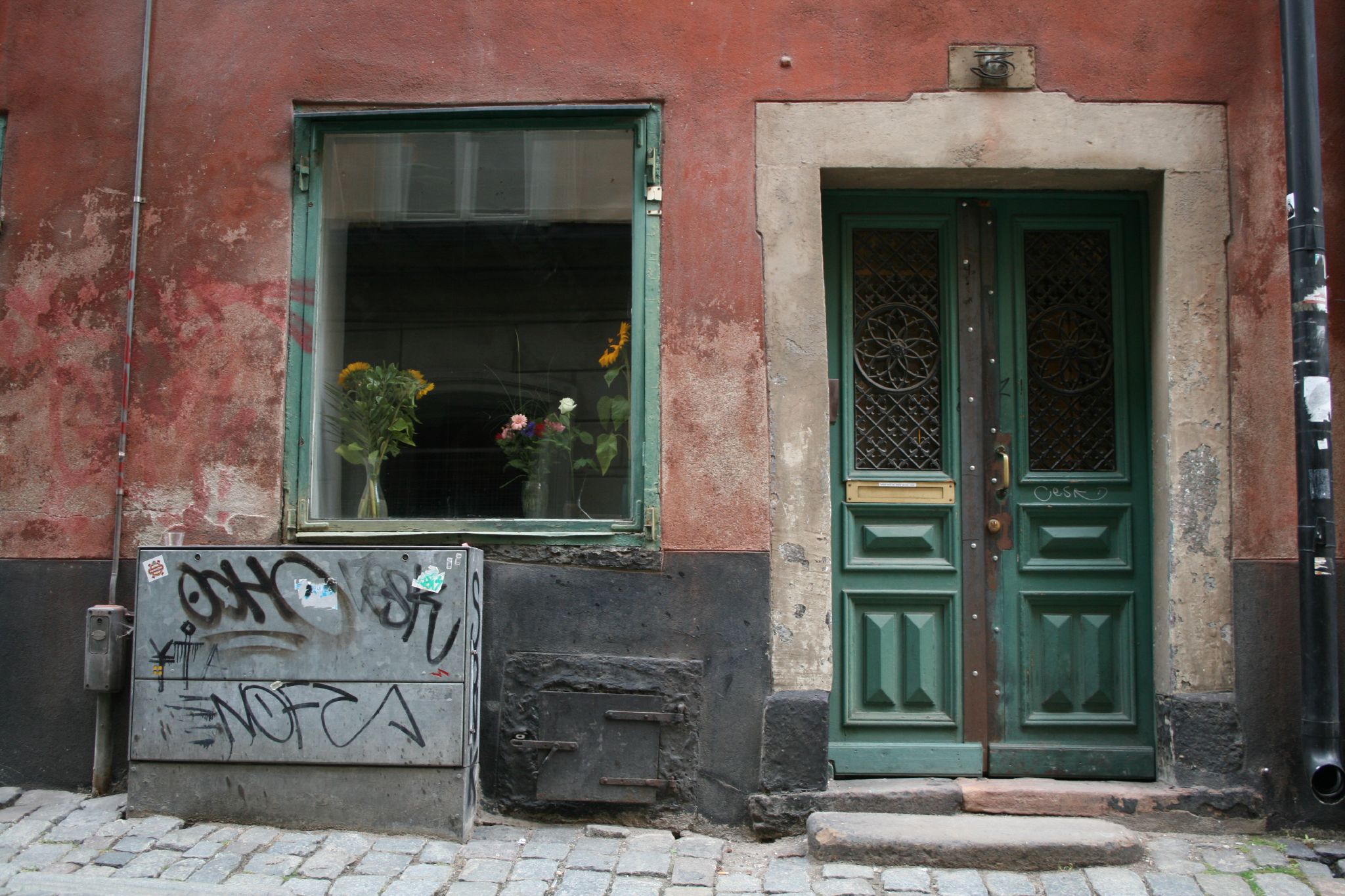 a red wall with green windows, flowers in the window and graffiti on the door