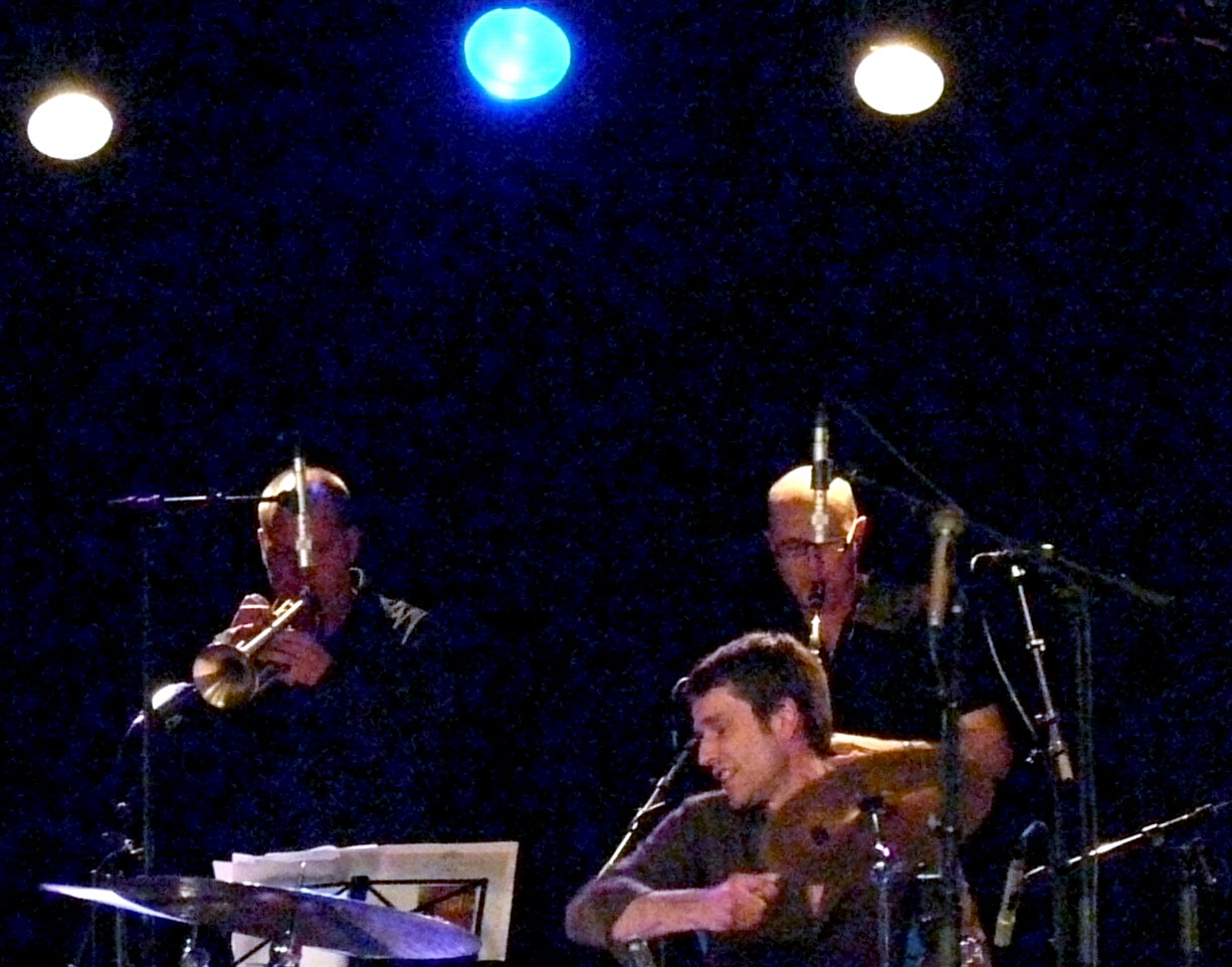 three men are standing in front of microphones, one is playing a musical instrument