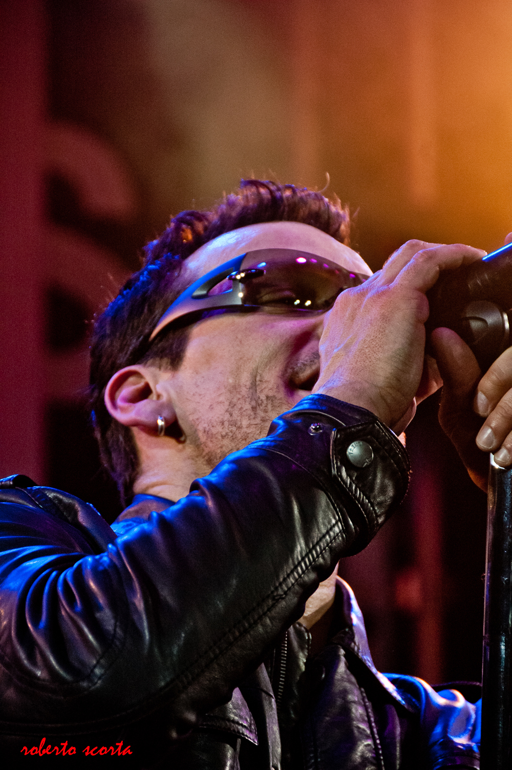 a man wearing a leather jacket and sunglasses, holds a microphone and covers the mouth with his hand