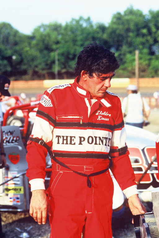 a man in racing gear standing near other cars