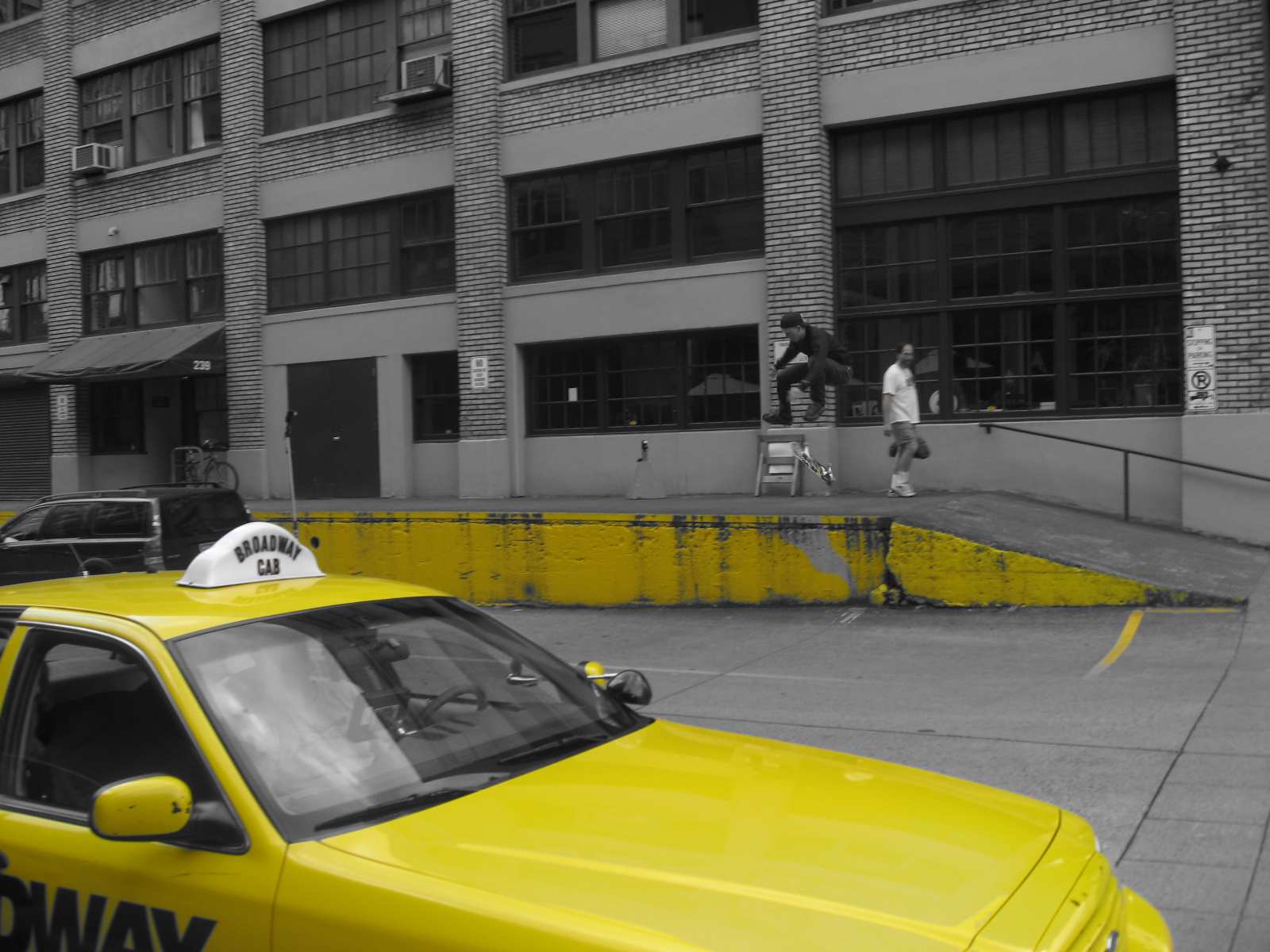 two people skateboard across a parking lot, next to a yellow taxi
