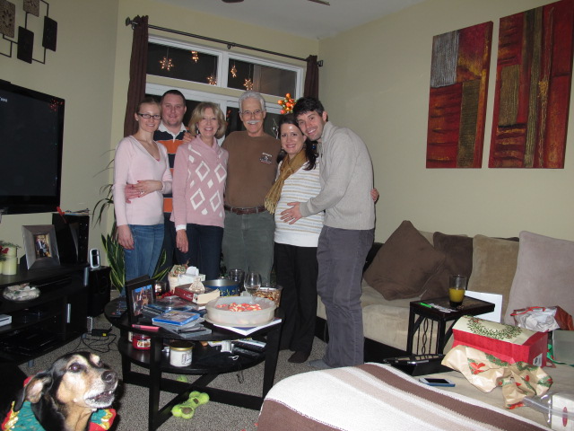 four people and a dog standing in front of a cake in a living room