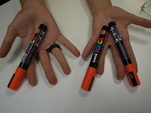 two hands, one with orange writing utensils and three other with black writing writing pens