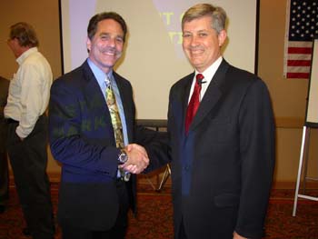 two men in suits shake hands in front of a projector screen
