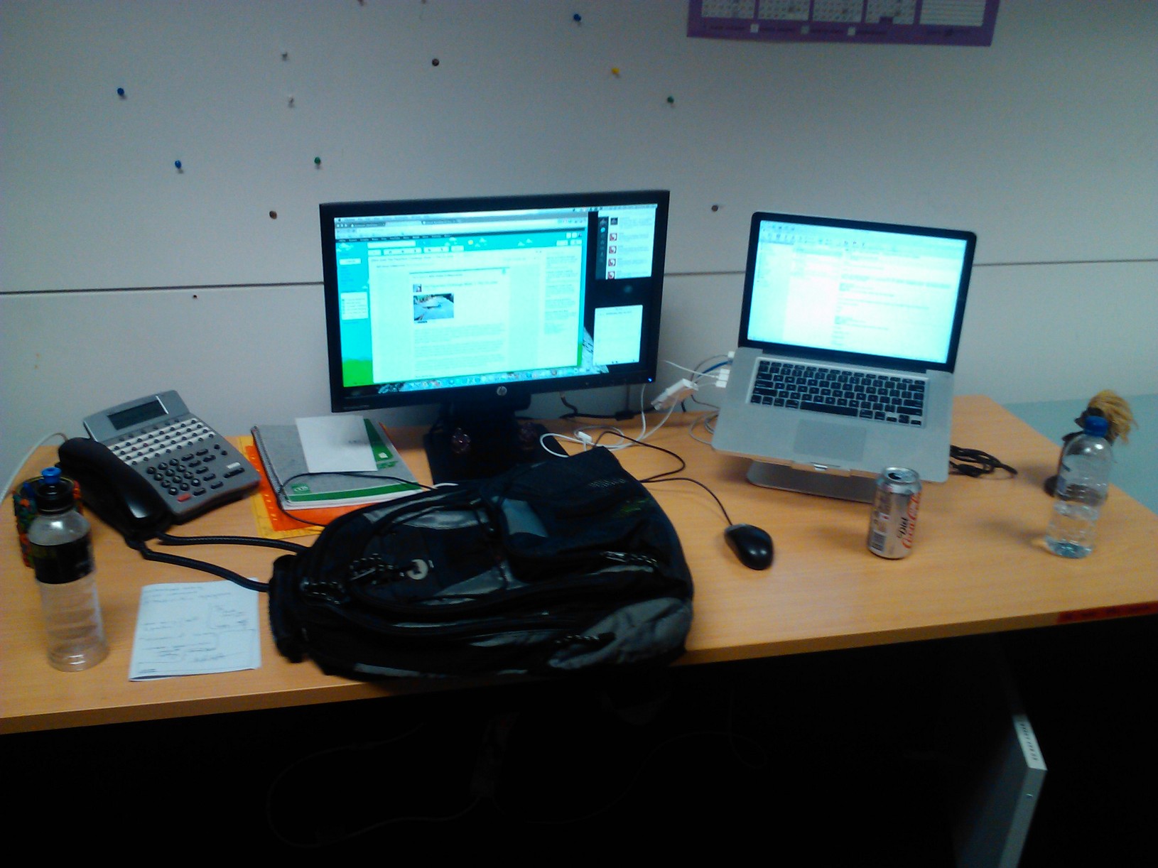 three computers are on a desk with a bag, telephone, laptop and cell phone