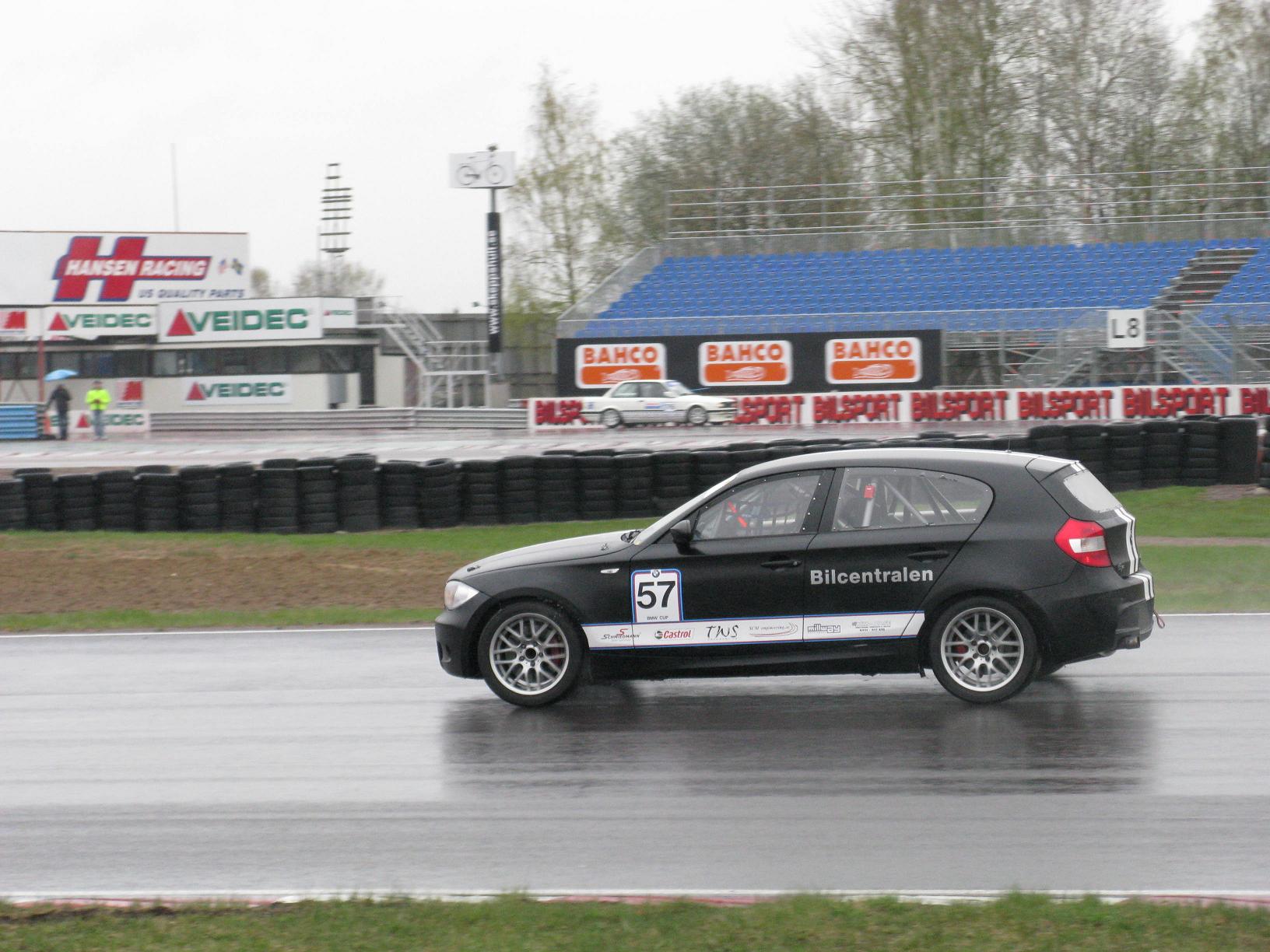 a car driving on a wet track with a sign for the race