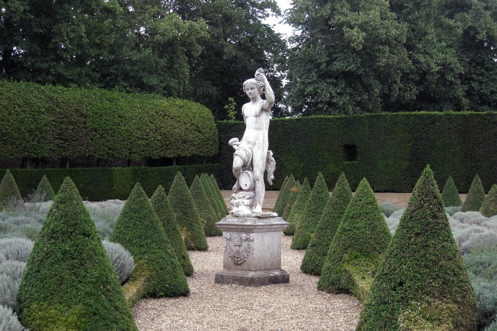a statue in front of many hedges, shrubs and a birdbath
