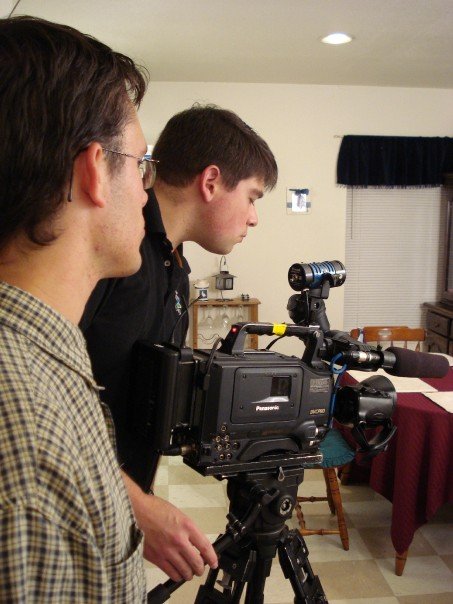 two men stand behind a camera on a tripod