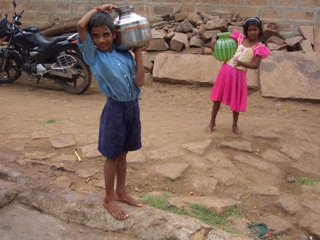 two little girls standing near rocks and one carrying a metal bowl