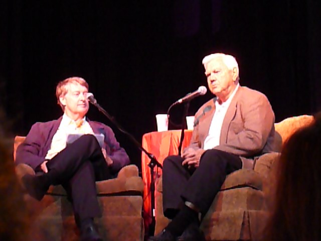 two men sit side by side on a stage while one speaks into a microphone
