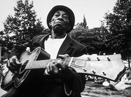 an older man with a hat sitting and playing a guitar