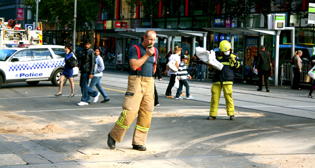 people walk in the street as a fireman talks on his cell phone