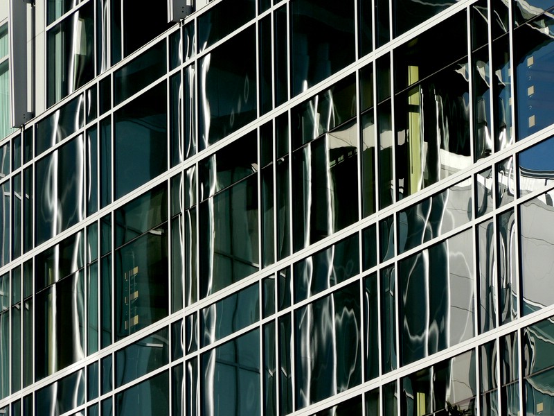 a clock stands near some glass windows and is reflecting a building