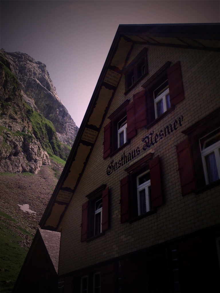 an old building with open shutters on the windows, stands below a huge rock face