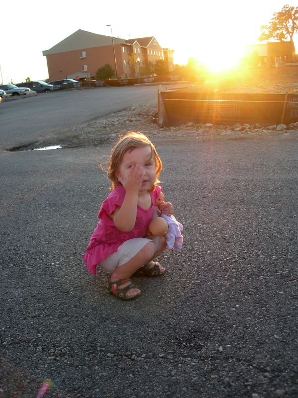 the little girl is kneeling on her knees in a parking lot with the sun setting behind her