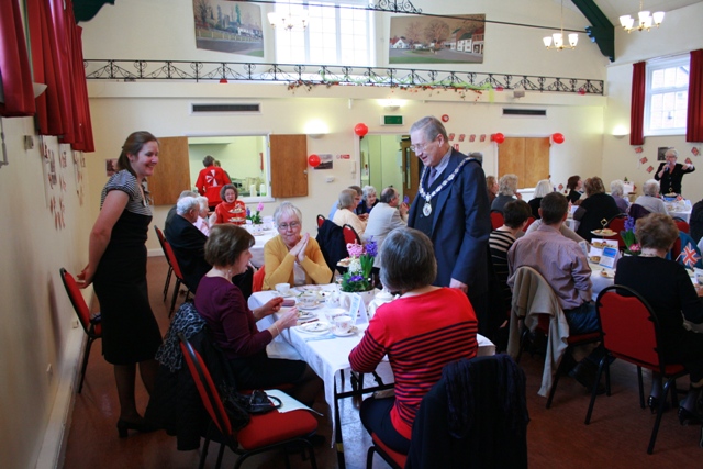 elderly people sit at long tables in a room with red curtains