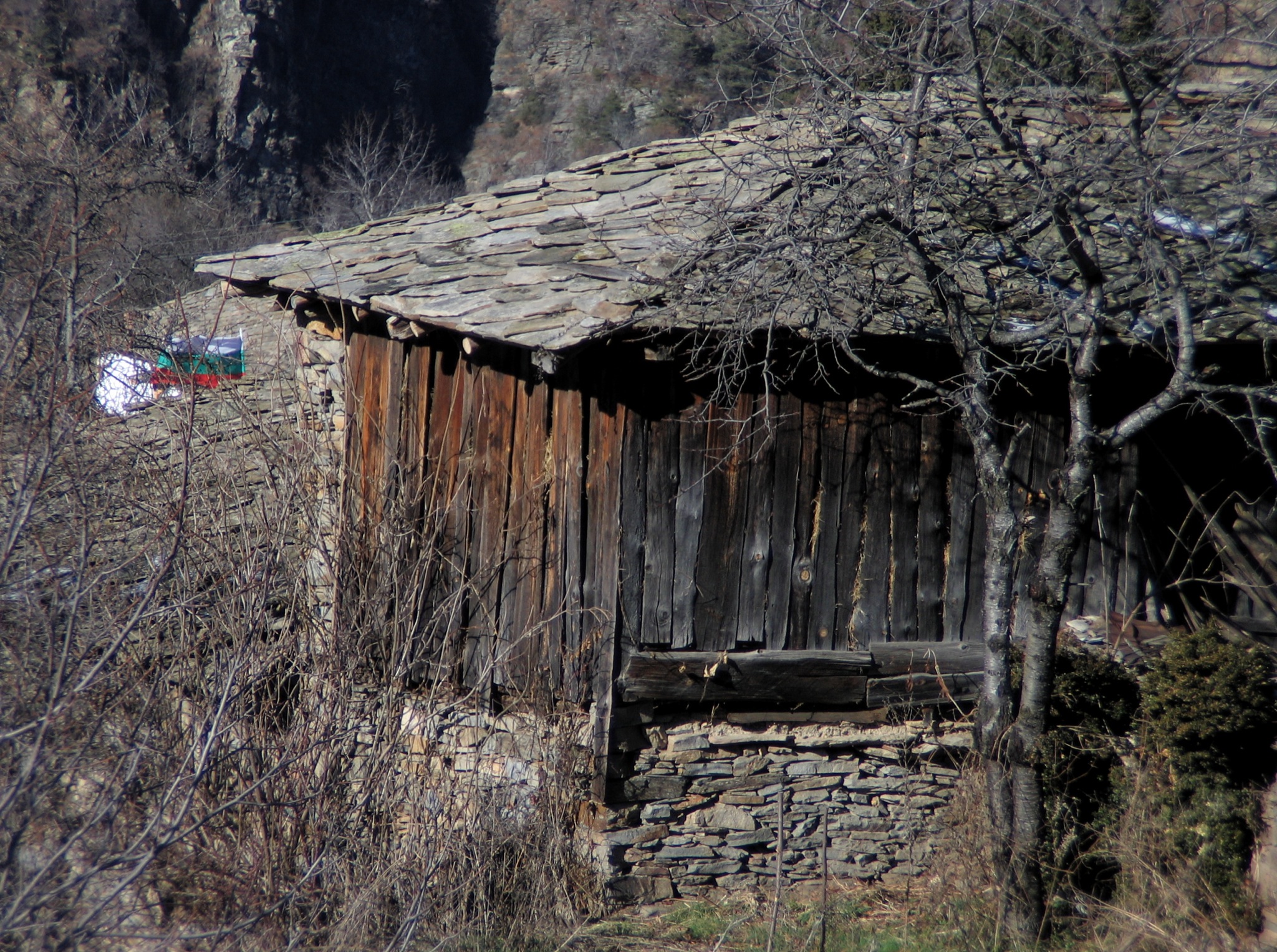 an old barn with a stone wall and a wooden roof and a few birds flying around