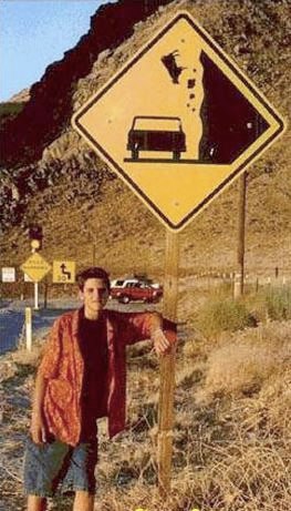a young man leaning on a sign next to some mountains