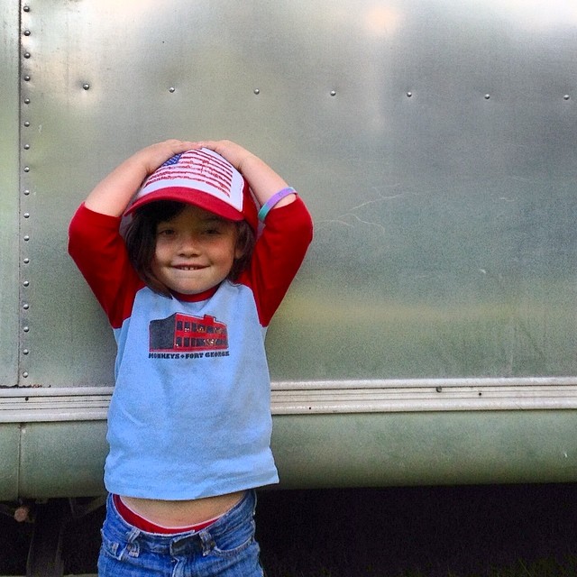 a boy standing in front of a silver truck wearing a blue shirt and red hat