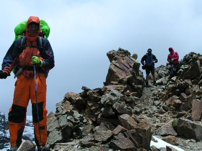 man wearing orange overalls standing on rocky mountain with backpack