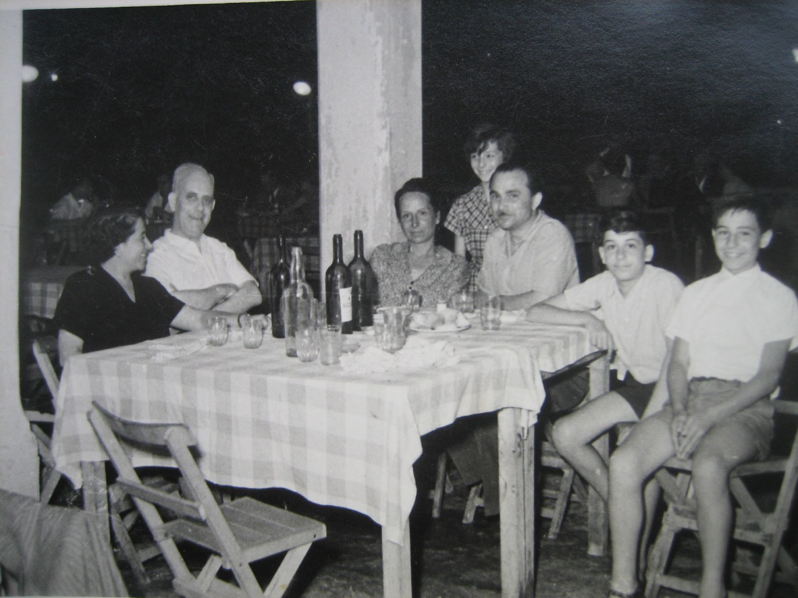 a family with four young adults are seated at a long table, eating food and drinking wine