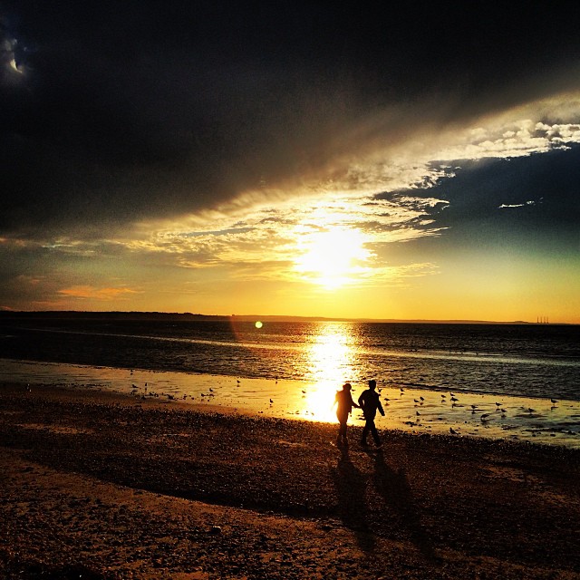 two people are walking along the beach as the sun sets