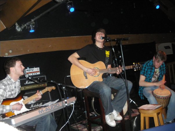 three musicians playing instruments and singing while sitting on stools