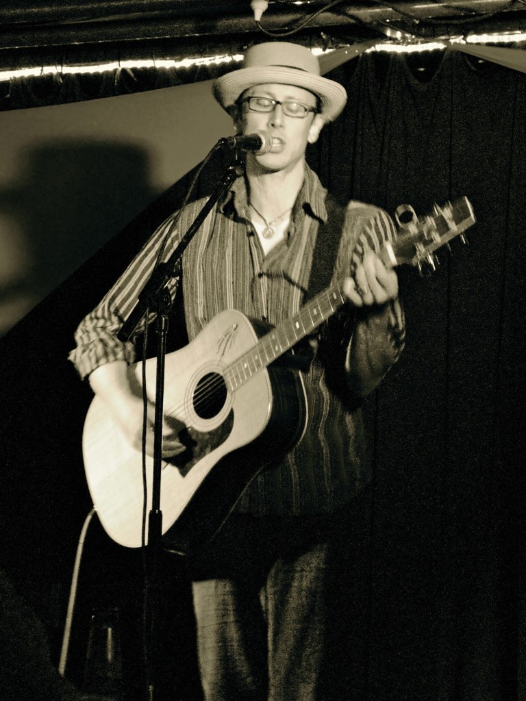 man wearing glasses playing a guitar and singing into microphone