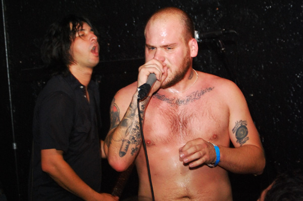 shirtless man singing into a microphone while a bearded man looks on